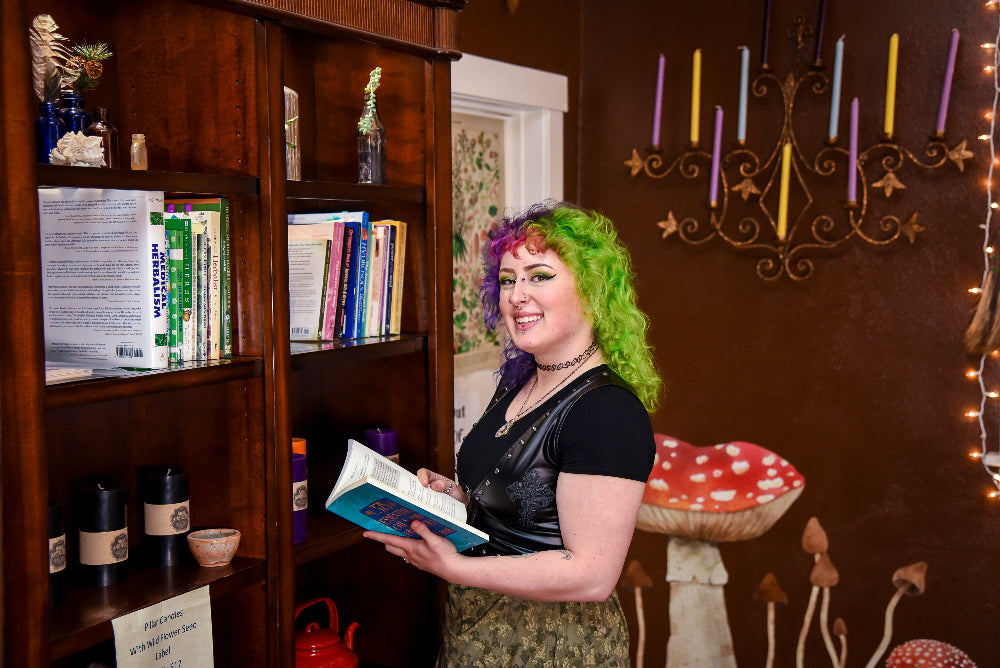 Person with colorful hair holding a book in a local herb shop with bookshelves and decorative lights.